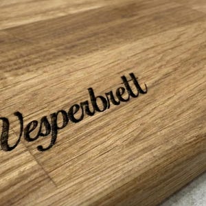 May include: A wooden cutting board with the word "Vesperbrett" engraved in a stylish font. The board has a natural wood grain pattern and a decorative element etched in the upper right corner.