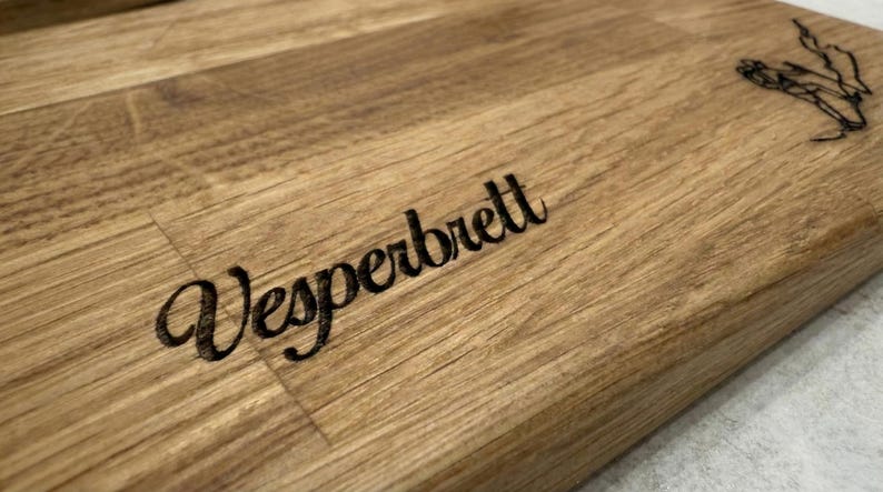 May include: A wooden cutting board with the word "Vesperbrett" engraved in a stylish font. The board has a natural wood grain pattern and a decorative element etched in the upper right corner.