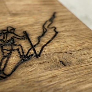 May include: Close-up of a wooden surface with a black etched design of a person hiking up a mountain. The hiker is wearing a backpack and using trekking poles. The wood has a natural grain and a warm, brown color.