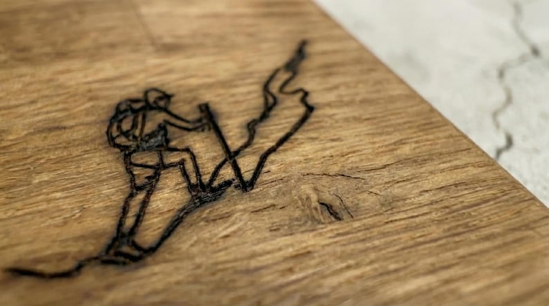 May include: Close-up of a wooden surface with a black etched design of a person hiking up a mountain. The hiker is wearing a backpack and using trekking poles. The wood has a natural grain and a warm, brown color.
