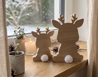 May include: Two light-colored wooden reindeer silhouettes of different sizes, each featuring a small white pom-pom tail. The figures are positioned on a wooden surface alongside a small potted plant and a lit candle in a glass holder.