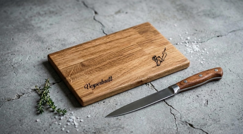 May include: A rectangular wooden cutting board with the word "Vesperbrett" engraved on it, along with a small illustration. A stainless steel knife with a wooden handle rests beside the board. Thyme sprigs and salt are also present.