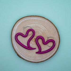 Könnte beinhalten: Zwei rosafarbene herzförmige Ohrringe mit silbernen Ringen. Die Ohrringe liegen auf einer Holzoberfläche.