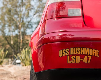 May include: A red car with a gold sticker that says "USS RUSHMORE LSD-47" on the back bumper.