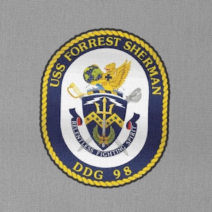 Puede incluir: Un parche bordado azul y dorado con el texto "USS Forrest Sherman DDG 98" y un escudo con un tridente, una espada y un pájaro.