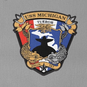 以下が含まれることがあります： 金色の縁取りと白い文字で「USS MICHIGAN TUEBOR」と書かれた紺色のパッチ。パッチには、潜水艦のシルエットと両側に魚が描かれた盾が描かれている。盾は赤、白、青の旗で囲まれている。「SSGN 727」という文字が盾の下に印刷されている。