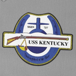 May include: A blue and yellow embroidered patch with a white background. The patch features a horseshoe, a cross, a gun, and the text "USS KENTUCKY" and "Thoroughbred of the Fleet".