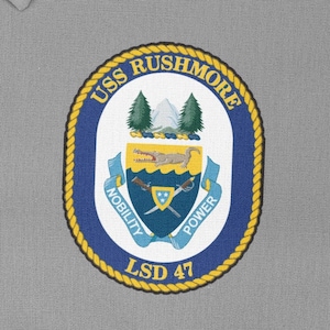 May include: A blue and gold circular patch with the text "USS RUSHMORE LSD 47" and a crest featuring a mountain range, a crocodile, and crossed swords with the words "NOBILITY" and "POWER".