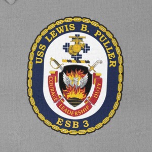 Könnte beinhalten: Ein marineblauer und goldener runder Patch mit dem Text "USS LEWIS B. PULLER ESB 3" und einem Wappen mit einem Schild, auf dem sich ein Phönix, Schwerter und die Worte "Mut, Führung, Pflicht" befinden.