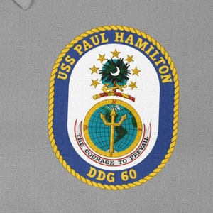 Può includere: Una camicia grigia con una toppa ricamata blu navy e oro. La toppa presenta il testo "USS PAUL HAMILTON DDG 60" e un emblema con un globo, un tridente e delle stelle. È inclusa anche la frase "THE COURAGE TO PREVAIL".
