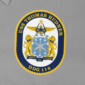 May include: A navy blue and gold embroidered patch with the text "USS THOMAS HUDNER DDG 116" and a crest featuring a ship, two lions, and the text "ABOVE ALL OTHERS".