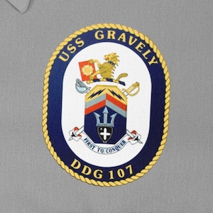 May include: A grey shirt with a navy blue and gold embroidered patch. The patch displays the text "USS GRAVELY" and "DDG 107", alongside a crest with a trident, sword, and the words "FIRST TO CONQUER".