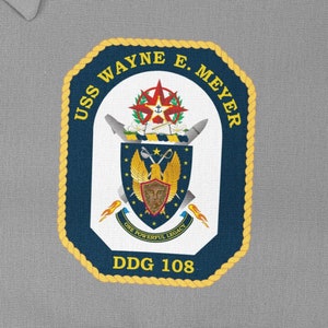 Può includere: Una toppa blu navy con bordatura in corda dorata e il testo "USS WAYNE E. MEYER DDG 108". La toppa presenta uno stemma con un'aquila dorata, uno scudo con un viso e una stella con una rosa dei venti.