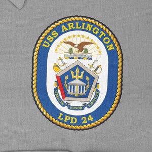Könnte beinhalten: Ein blauer und goldener gestickter Aufnäher mit dem Text "USS ARLINGTON LPD 24" und einem Wappen mit einem Gebäude, einem Dreizack und einem Vogel.