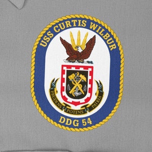 May include: A navy blue and gold embroidered patch with the text "USS CURTIS WILBUR DDG 54" and a crest featuring an eagle, crossed hammers, and an anchor.