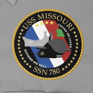 Pode incluir: Um remendo circular preto e dourado com o texto "USS MISSOURI" e "SSN 780" em uma borda de corda dourada. O remendo apresenta um fundo vermelho, branco e azul com uma silhueta de um submarino e um encouraçado. O texto "UNITED WE STAND, DIVIDED WE FALL" também está incluído.