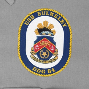 Può includere: Una toppa circolare blu navy e oro con il testo "USS BULKELEY DDG 84" e uno stemma con uno scudo con un'ancora e una stella.
