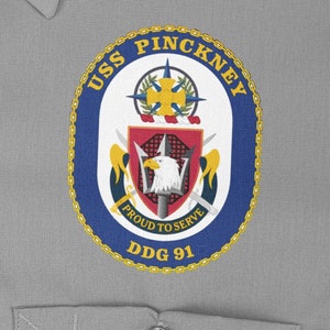 Può includere: Una toppa circolare blu navy e oro con il testo "USS PINCKNEY DDG 91" e le parole "Proud to Serve" all'interno di uno scudo con un'aquila calva.