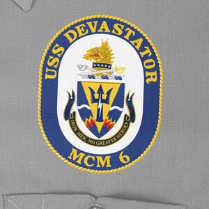 May include: A navy blue and gold circular patch with the text "USS DEVASTATOR MCM 6" and a crest featuring a trident, a flame, and the text "Iron Men. No Greater Heroes."
