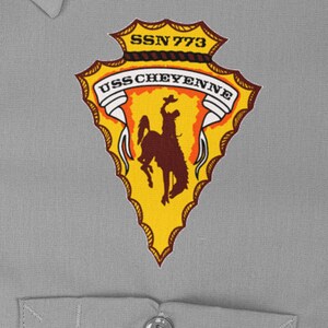 May include: A yellow and orange arrowhead patch with a brown silhouette of a cowboy riding a horse. The patch has the text "USS CHEYENNE" and "SSN 773" on it.