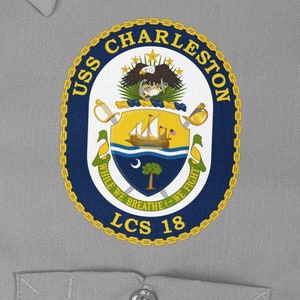 May include: A blue and gold circular patch with the text "USS CHARLESTON LCS 18" and a crest featuring a ship, a palmetto tree, and a crescent moon. The text "WHILE WE BREATHE WE FIGHT" is wrapped around the crest.