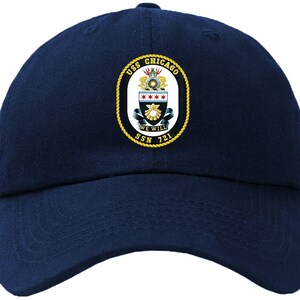 Può includere: Un cappello da baseball blu navy con una toppa ricamata in oro che presenta lo stemma dell'USS Chicago SSN 721. Lo stemma include le parole "USS Chicago" e "We Will" con uno scudo e stelle.