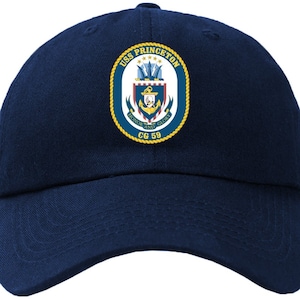 Könnte beinhalten: Eine marineblaue Baseballkappe mit einem goldenen gestickten Aufnäher, der das Abzeichen der USS Princeton zeigt, einschließlich des Schiffsnamens, fünf Sternen, einem Anker und den Worten "Ehre und Ruhm" mit der Schiffsbezeichnung "CG 59" darunter.