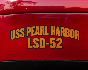 May include: Close-up of the rear of a red car with a black bumper. A sticker on the car reads "USS PEARL HARBOR LSD-52" in gold lettering. The car's taillight is visible.