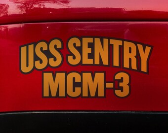 May include: A red car with a gold sticker that says "USS SENTRY MCM-3" on the back bumper.