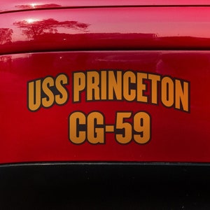 Könnte beinhalten: Ein rotes Auto mit einem goldenen Aufkleber, auf dem "USS PRINCETON CG-59" steht, auf der hinteren Stoßstange.