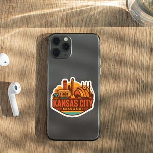 a smartphone with a sticker of Kansas City Missouri on its back, along with a pair of white earbuds and a glass of water on a wooden surface.