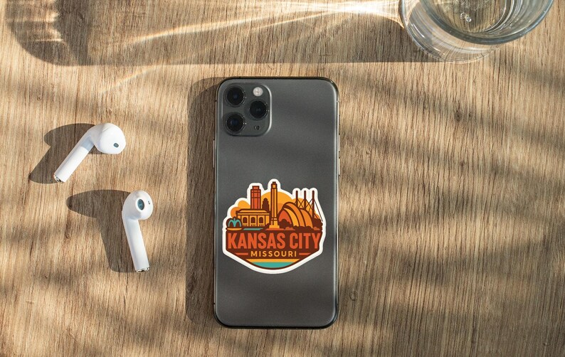 a smartphone with a sticker of Kansas City Missouri on its back, along with a pair of white earbuds and a glass of water on a wooden surface.