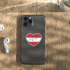 May include: A black iPhone with a white heart sticker featuring the Austrian flag on the back. The phone is on a wooden surface with two white wireless earbuds.