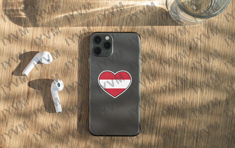 May include: A black iPhone with a white heart sticker featuring the Austrian flag on the back. The phone is on a wooden surface with two white wireless earbuds.