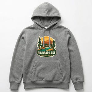 May include: A gray hooded sweatshirt with a front pocket and drawstring. The sweatshirt features a colorful graphic of Big Bear Lake, California, with trees, a lake, and a sunset. The text "BIG BEAR LAKE CALIFORNIA" is below the graphic.