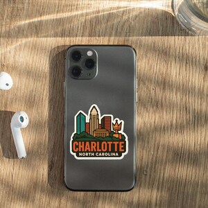 a smartphone with a sticker of the Charlotte, North Carolina skyline, along with a pair of white earbuds and a glass of water on a wooden surface.