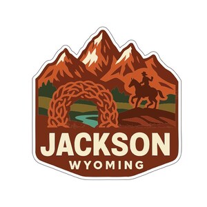 Könnte beinhalten: Ein Aufkleber mit einer Bergkette, einem Cowboy auf einem Pferd und einem Torbogen über einem Fluss. Der Text "JACKSON WYOMING" wird in fetter Schrift angezeigt, mit einem braunen und orangen Farbschema.