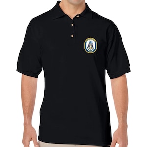 Könnte beinhalten: Schwarzes Poloshirt mit einem marineblauen und goldenen Emblem auf der linken Brust. Das Emblem zeigt ein Wappen mit Text und einem nautischen Design. Das Shirt hat einen Kragen und drei Knöpfe.