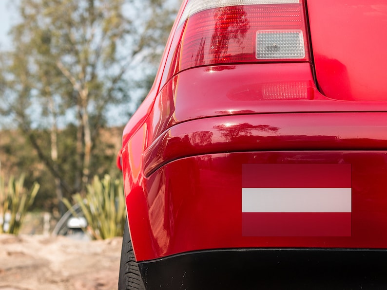 K&ouml;nnte beinhalten: Ein rotes Auto mit einem wei&szlig;en und roten Sto&szlig;f&auml;ngeraufkleber der lettischen Flagge.
