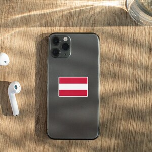 K&ouml;nnte beinhalten: Ein graues Smartphone mit einem wei&szlig;en und roten Aufkleber der lettischen Flagge auf der R&uuml;ckseite. Das Telefon liegt auf einer Holzoberfl&auml;che mit wei&szlig;en kabellosen Ohrh&ouml;rern.