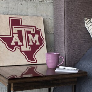 May include: A maroon and white canvas print of the state of Texas with the letters "TAM" inside the outline of the state. The print is sitting on a wooden end table next to a purple mug and a notebook.
