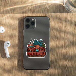 a smartphone with a sticker on it, placed on a wooden surface alongside a pair of white earbuds and a glass of water.
