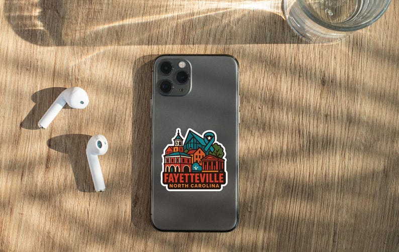 a smartphone with a sticker on it, placed on a wooden surface alongside a pair of white earbuds and a glass of water.