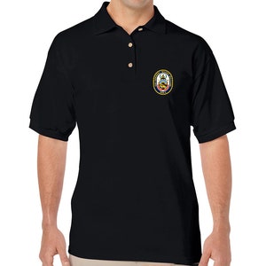 May include: Black polo shirt with a small embroidered patch on the left chest. The patch features a gold circle with a navy blue and white crest inside. The crest includes a ship, a lighthouse, and the text "Naval Weapons Station Seal Beach".