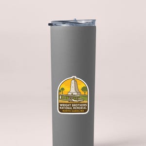 A gray insulated tumbler with a sticker on it, featuring an illustration of a monument and text that says "Wright Brothers National Memorial North Carolina".