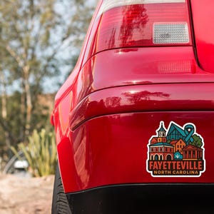 the rear end of a red car with a sticker on the trunk that features a colorful illustration of a cityscape.