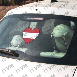 May include: A white car with a red heart sticker on the back window. The heart is decorated with the colors of the Austrian flag.