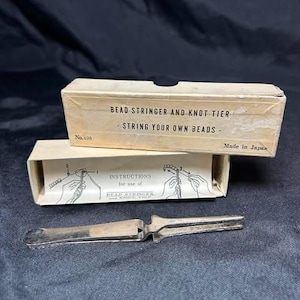 May include: A silver-colored metal bead stringer and knot tier tool with its original beige cardboard box. The box lid reads BEAD STRINGER AND KNOT TIER, STRING YOUR OWN BEADS, No. 498, and Made in Japan. The box interior shows line art instructions.
