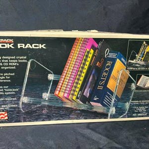 May include: A rectangular product box for a Super Rack Book Rack. The box features a clear acrylic book rack holding several books and manuals. Text on the box includes Super Rack Book Rack, Made in America, and assembled dimensions of 13 inches by 7 inches by 6 inches.