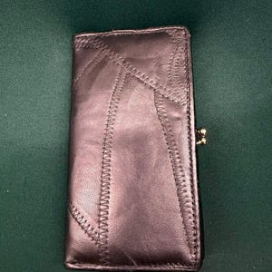 May include: A rectangular dark metallic leather wallet featuring a patchwork design with visible zigzag stitching along the seams. The wallet has a gold-toned metal kiss-lock clasp closure on the side, providing a secure way to keep the item closed.